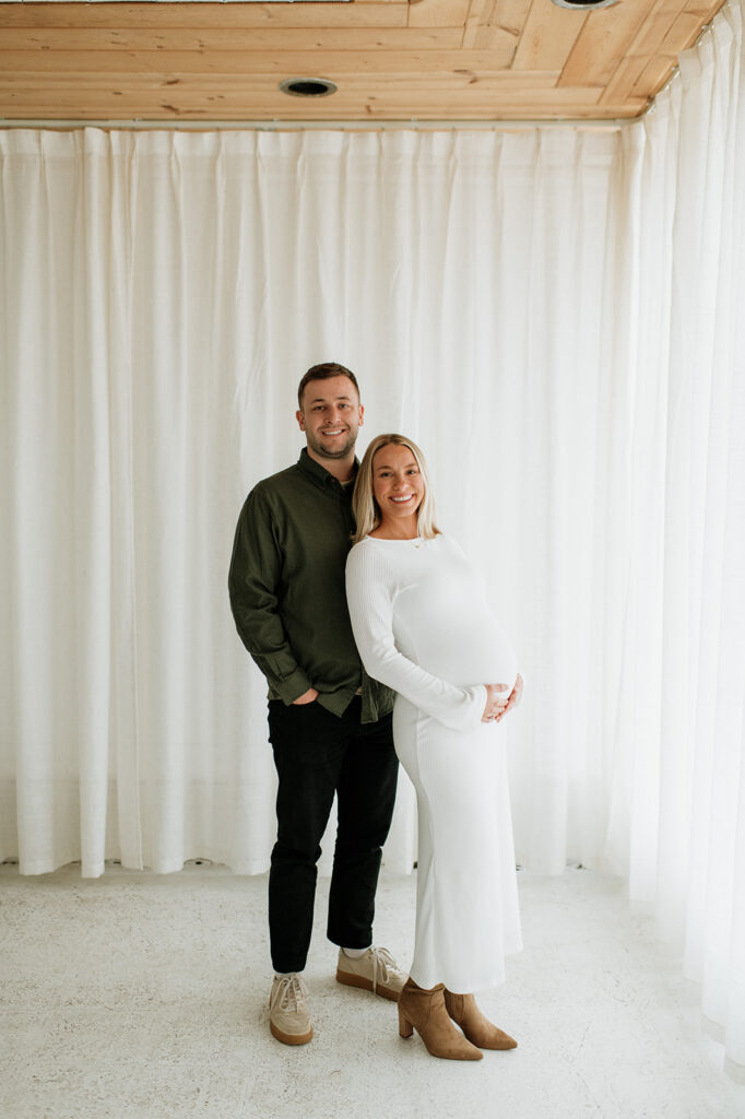 Pregnant woman in a white knit dress standing beside her partner during South Bend Indiana maternity photos in a bright studio.