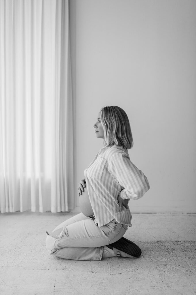 Black and white maternity portrait of a pregnant woman kneeling near a window in a bright studio for her South Bend, Indiana maternity photos.