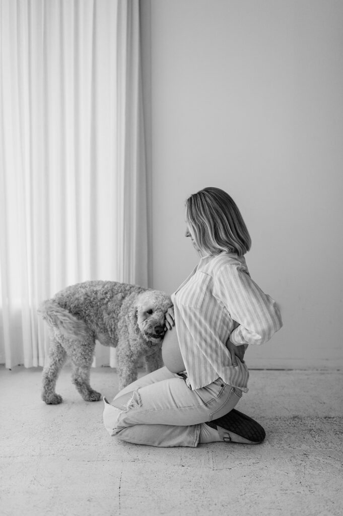 Black and white maternity photo of a pregnant woman kneeling as her dog stands beside her during her studio South Bend, Indiana maternity photos.