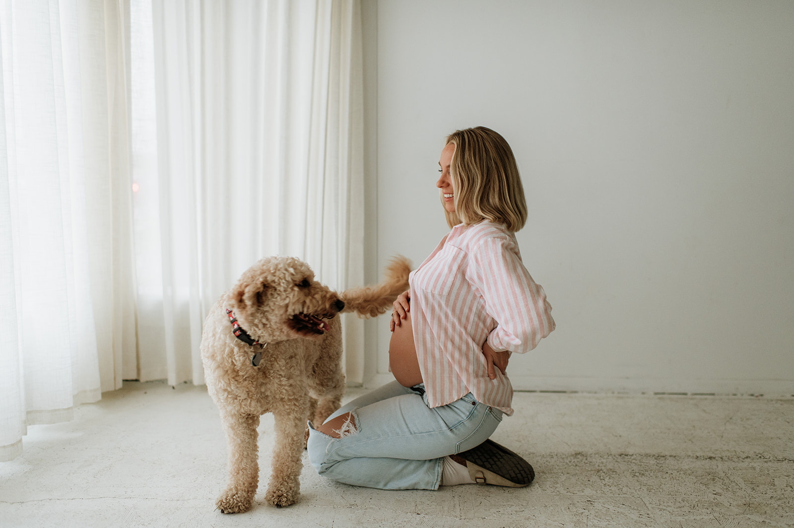 Pregnant woman kneeling on the studio floor while her dog stands nearby in a naturally lit space.