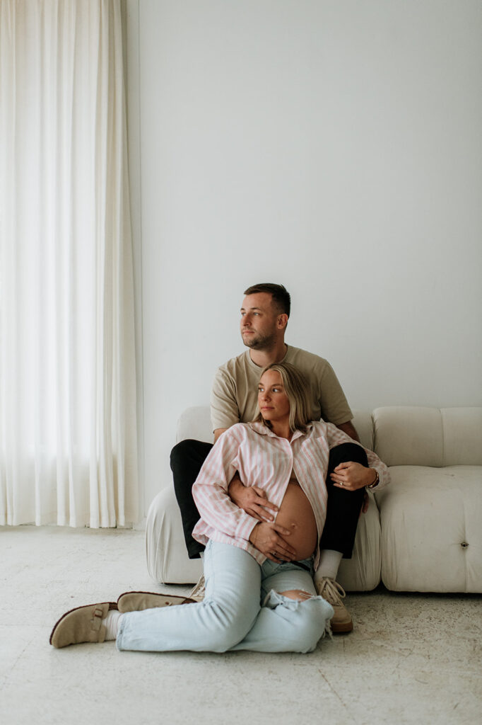 Expecting couple seated together on a white couch at Area Hall Art House during a clean, minimal South Bend, Indiana maternity session.