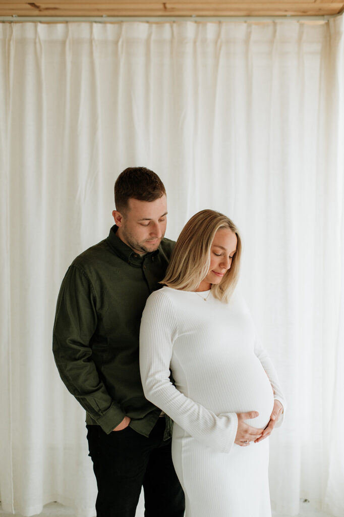 Pregnant woman and man looking down at her belly during South Bend Indiana maternity photos in a bright studio.