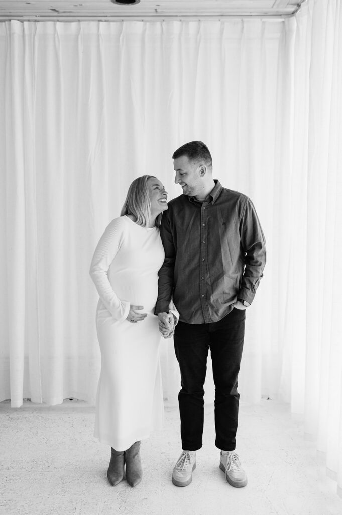 Expecting couple holding hands and smiling during South Bend Indiana maternity photos in a light-filled studio.