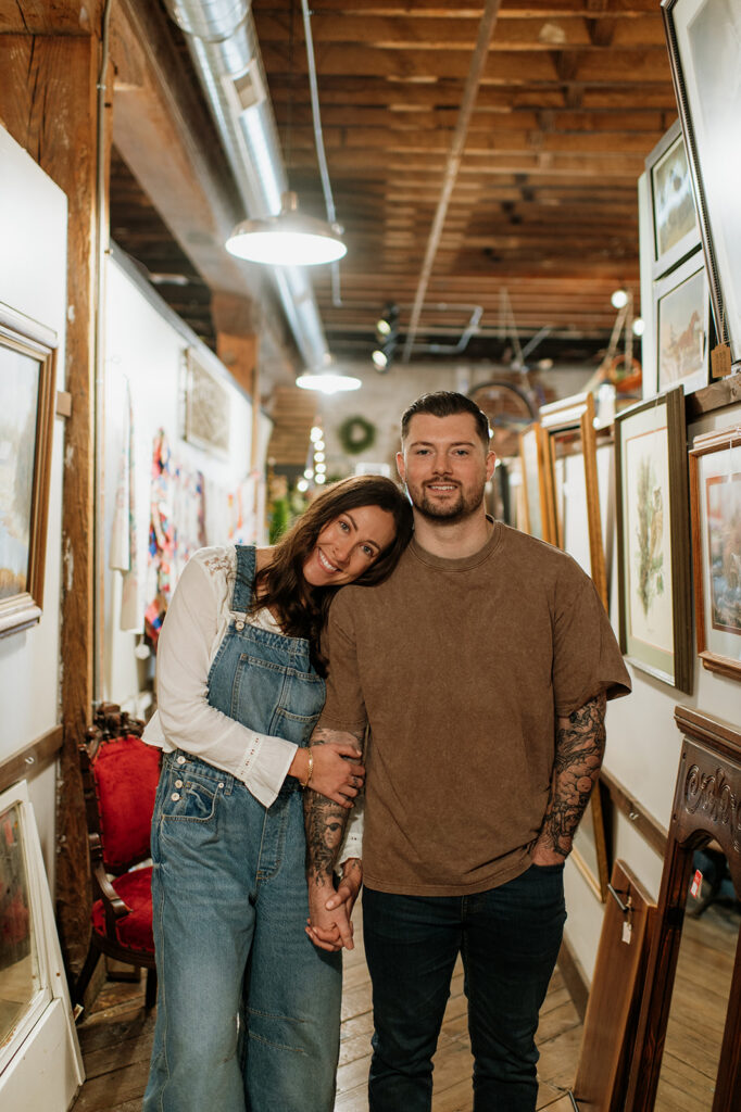 Couple posing in an antique shop holding hands during vintage style engagement photos.