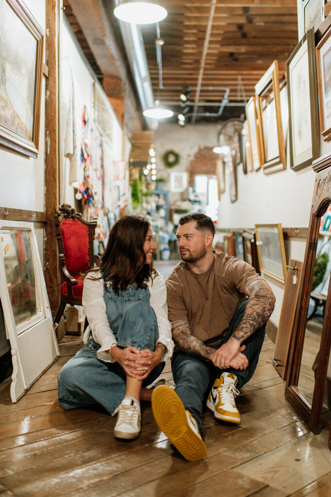 Smiling couple sitting on wooden floor surrounded by framed artwork during antique shop engagement session.