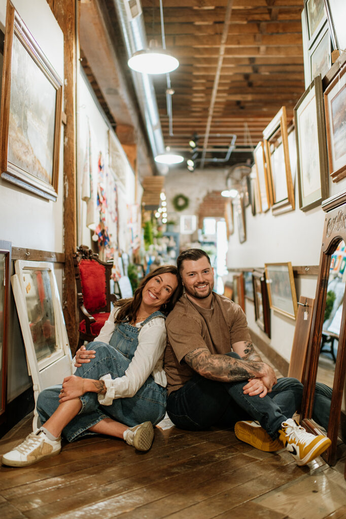 Smiling couple sitting on wooden floor surrounded by framed artwork during antique shop engagement session.