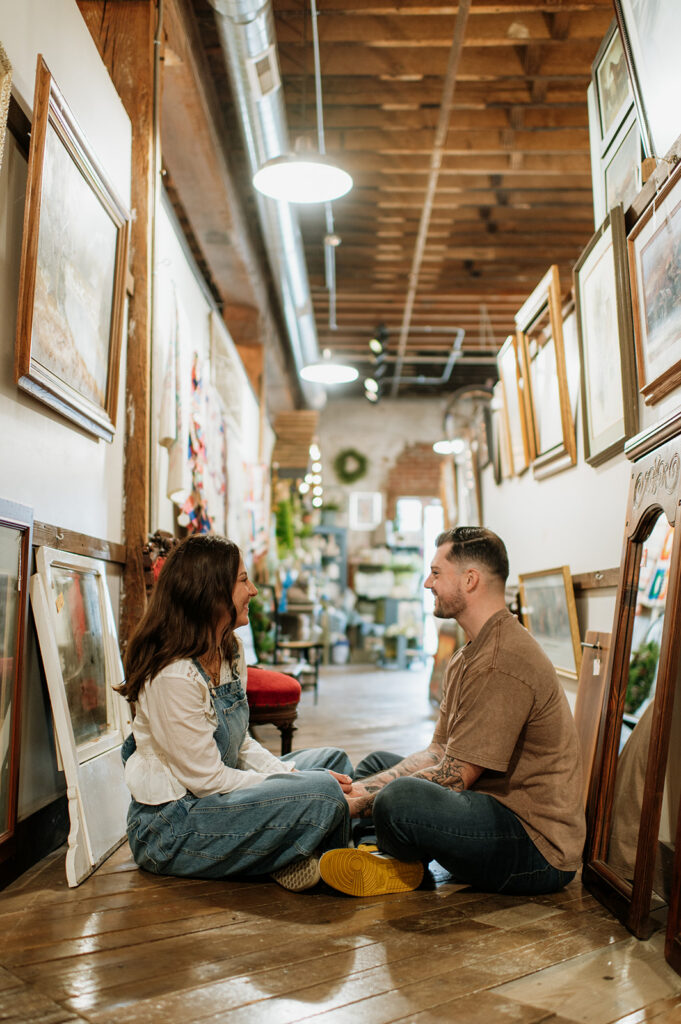 Smiling couple sitting on wooden floor surrounded by framed artwork during antique shop engagement session.