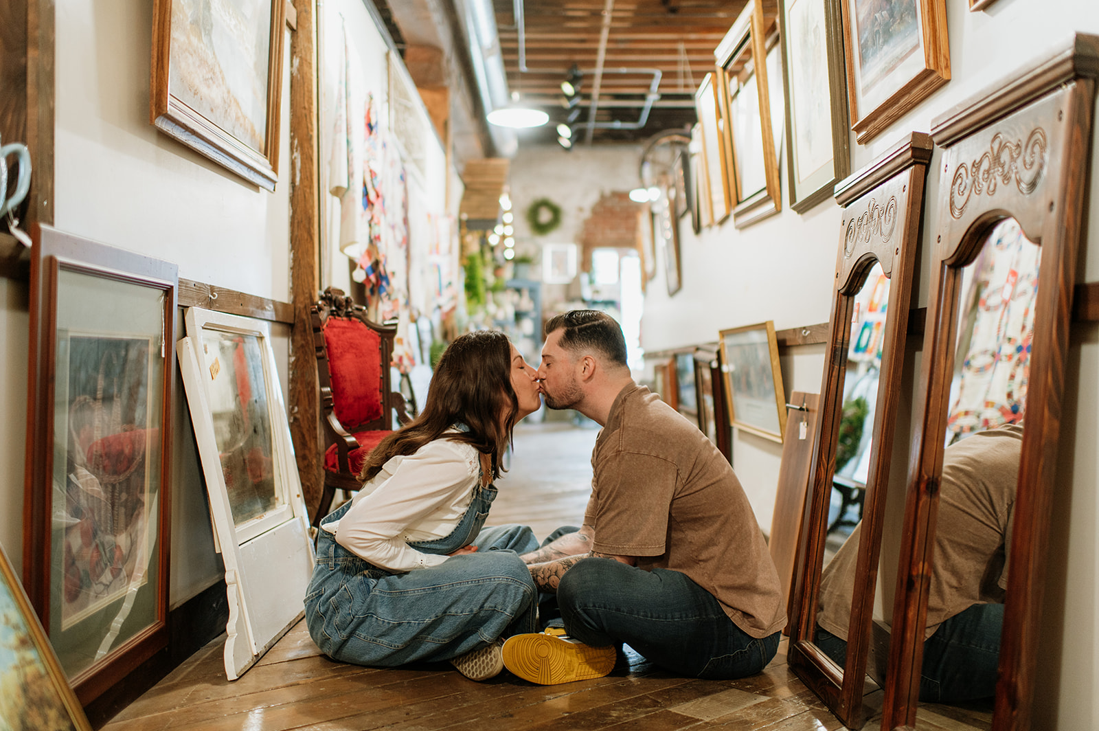 Kissing couple sitting on wooden floor surrounded by framed artwork during antique shop engagement session.