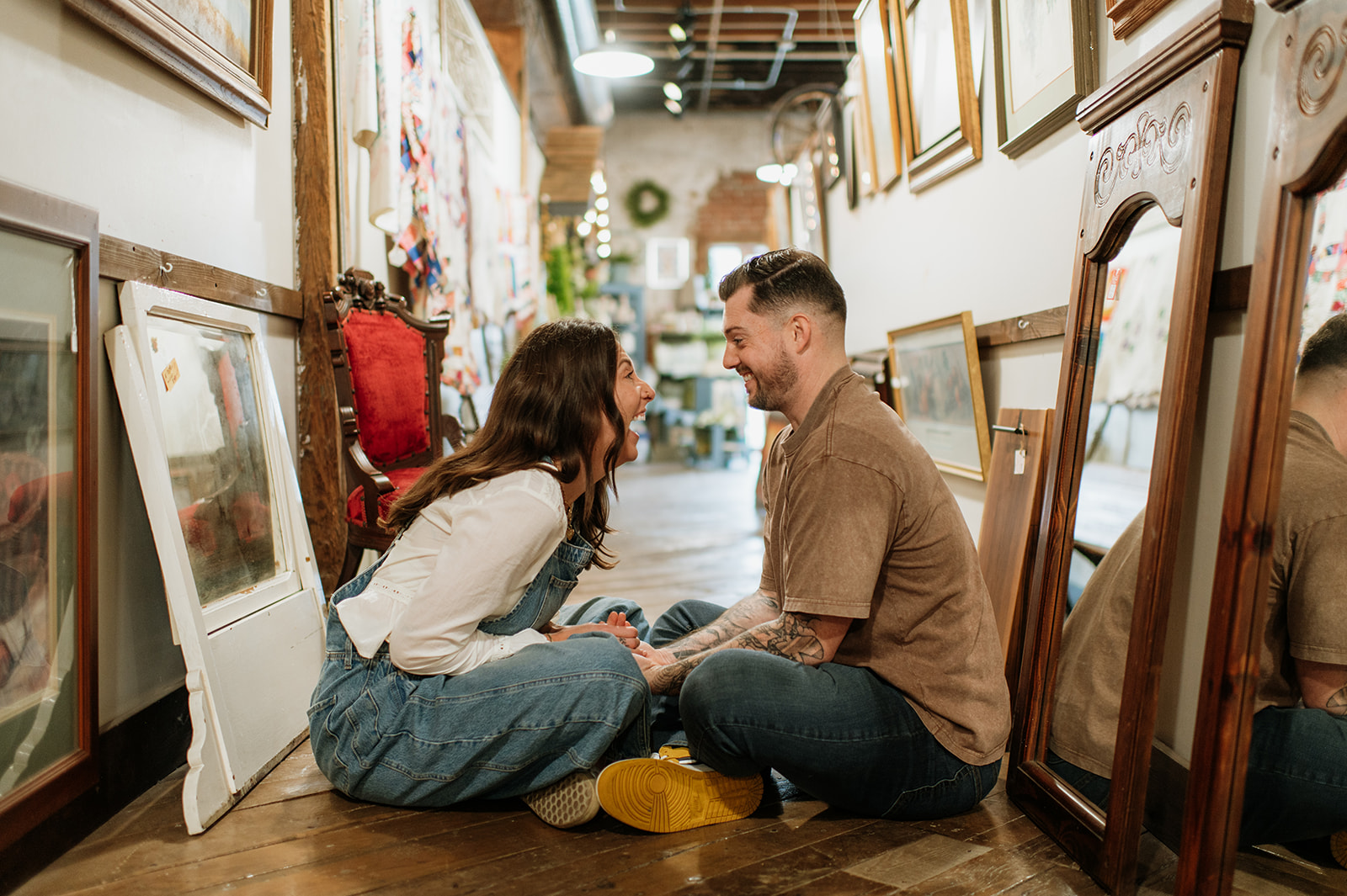 Couple laughing while sitting on wooden floor surrounded by framed artwork during antique shop engagement session.