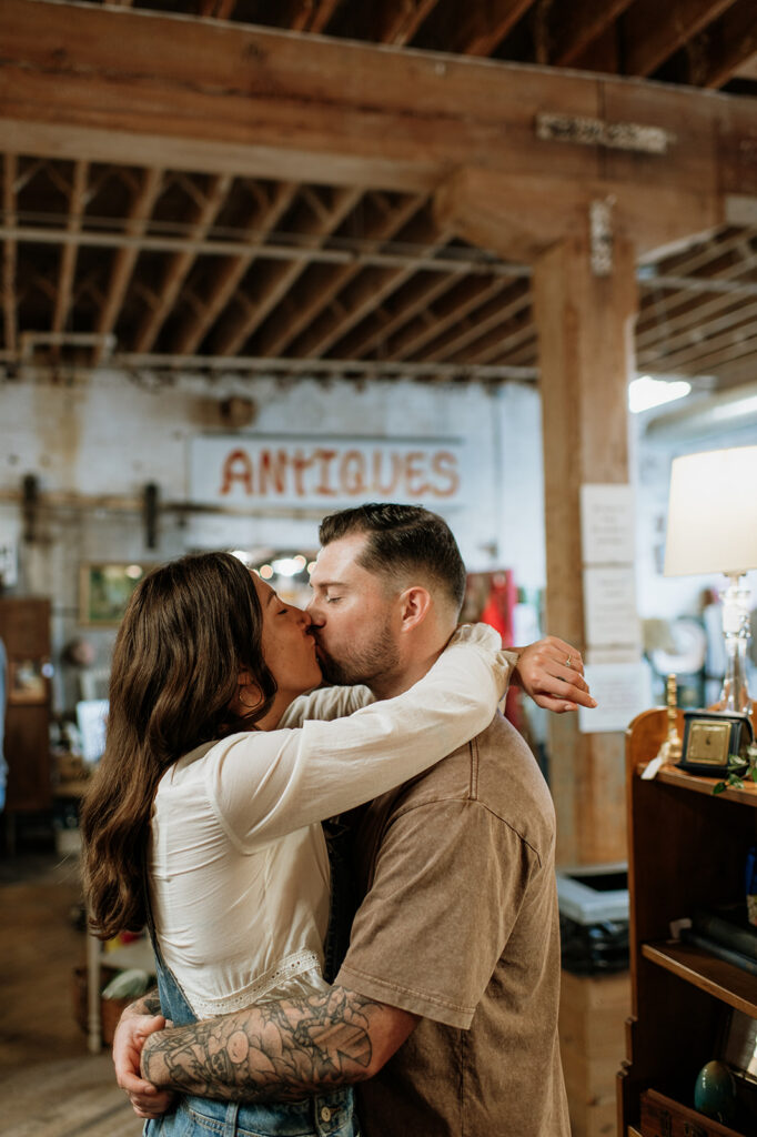 Couple kissing with an “antiques” sign during vintage style engagement photos near South Bend, Indiana.