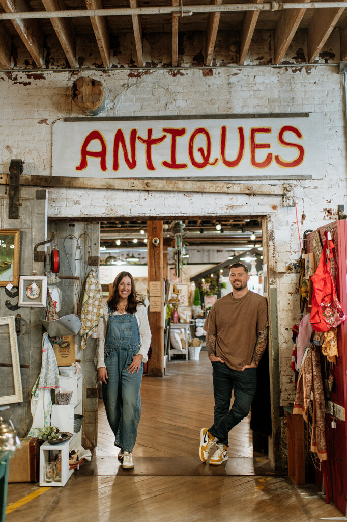 Couple standing in doorway under “antiques” sign during vintage style engagement photos.