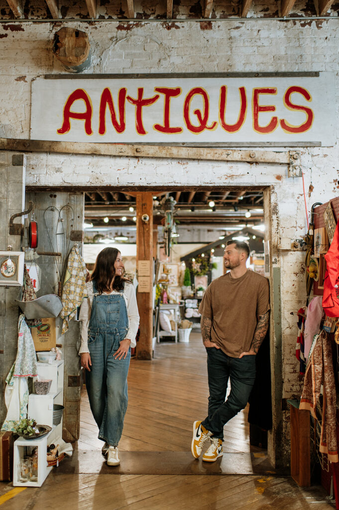 Couple standing in doorway under “antiques” sign during vintage style engagement photos.