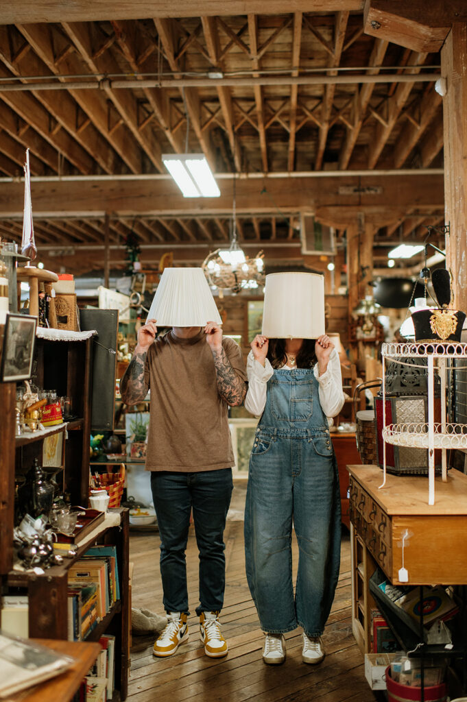 Playful couple holding lamp shades over their heads during antique shop engagement session.