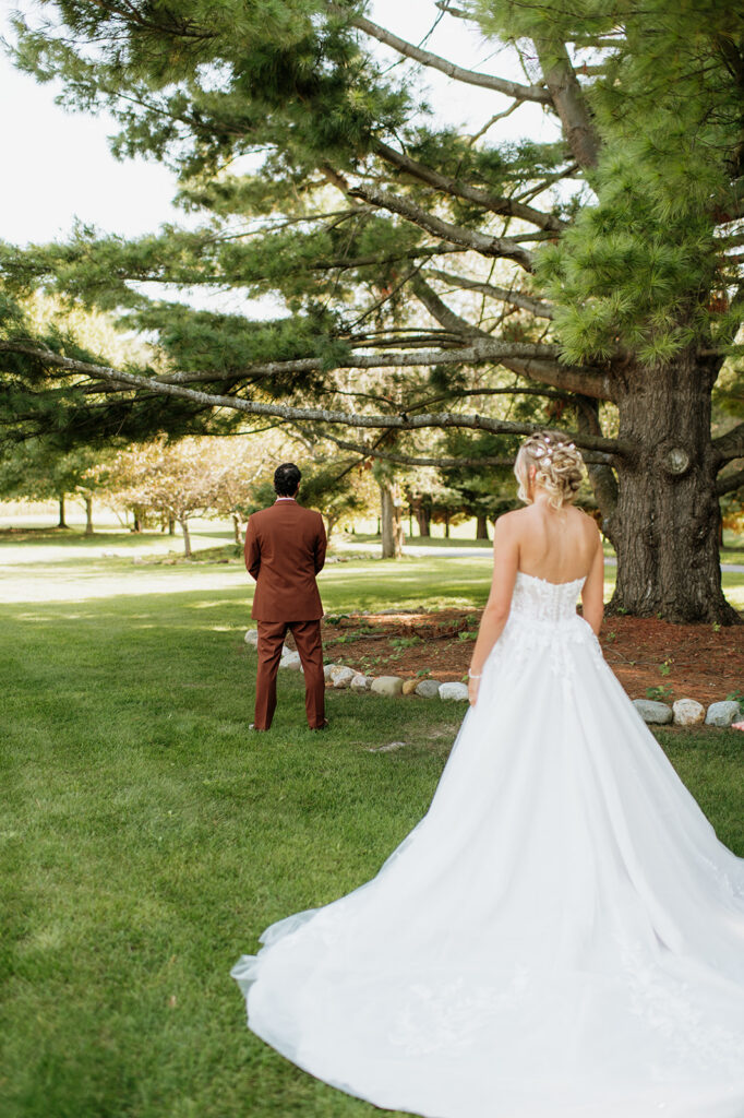Bride walking up behind the groom for their first look under tall pine trees at The Lodge at Rush Lakes wedding venue.