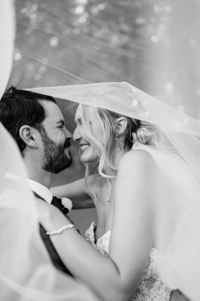 Close up black and white portrait of bride and groom smiling together under veil.