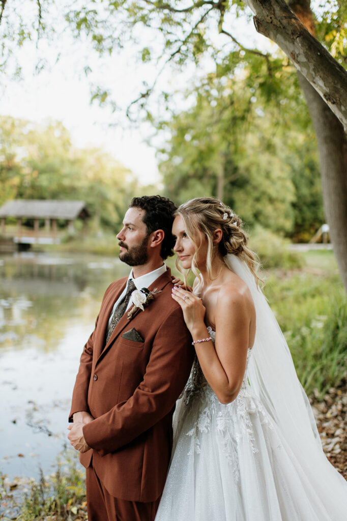 Bride and groom posing along lakeshore with trees and gazebo in the background.
