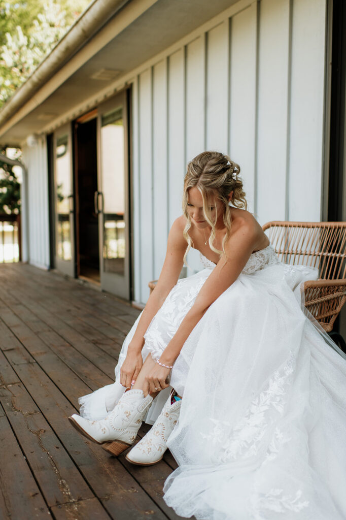Bride sitting on a wooden porch putting on white cowboy boots before her wedding at The Lodge at Rush Lakes.