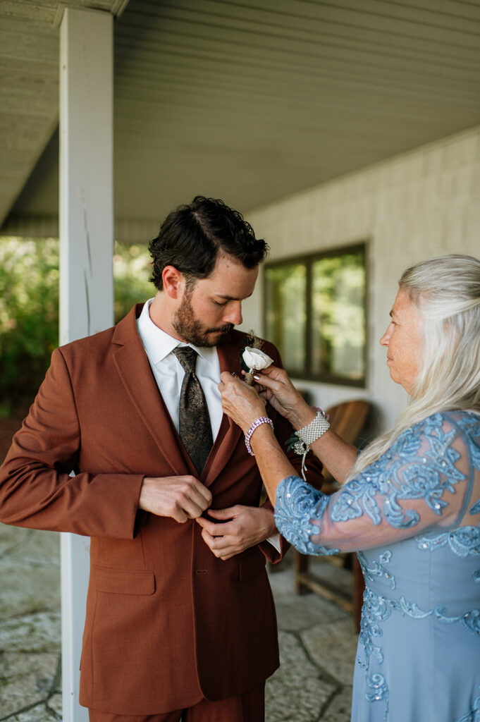 Groom getting ready in the guest house while his mother pins his boutonniere at The Lodge at Rush Lakes wedding venue.