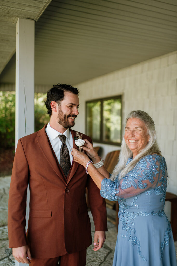 Groom smiling while his mother adjusts his boutonniere during getting ready at The Lodge at Rush Lakes wedding.