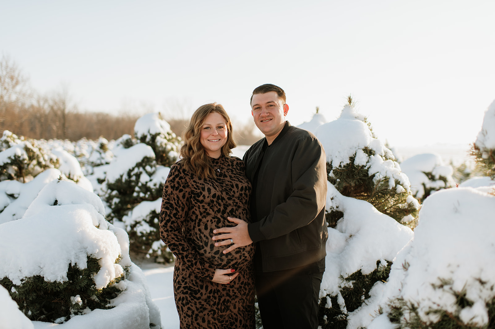 Couple standing together in a snowy pine tree farm during outdoor winter maternity photos in Northern Indiana.