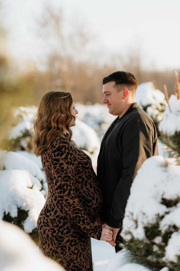 Expectant couple looking at each other between snow-covered evergreen trees during outdoor winter maternity photos.