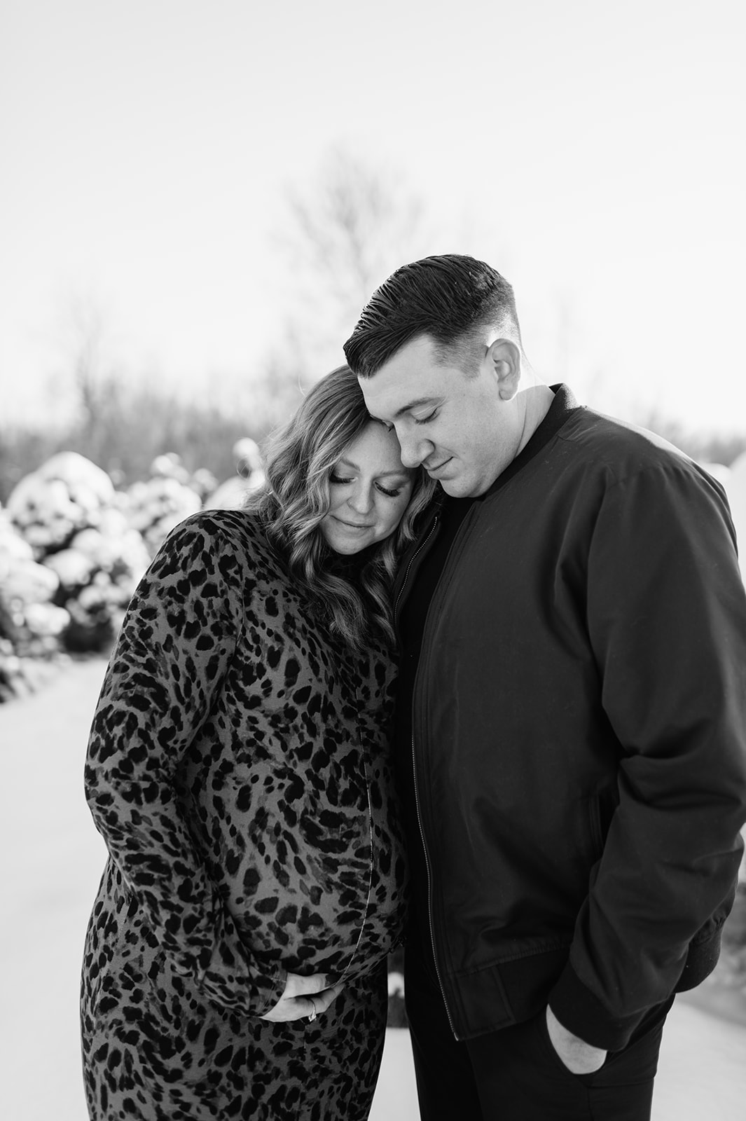 Black and white maternity portrait of couple with baby bump in a snowy tree farm.