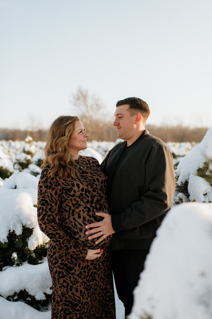Expectant couple looking at each other between snow-covered evergreen trees during outdoor winter maternity photos.