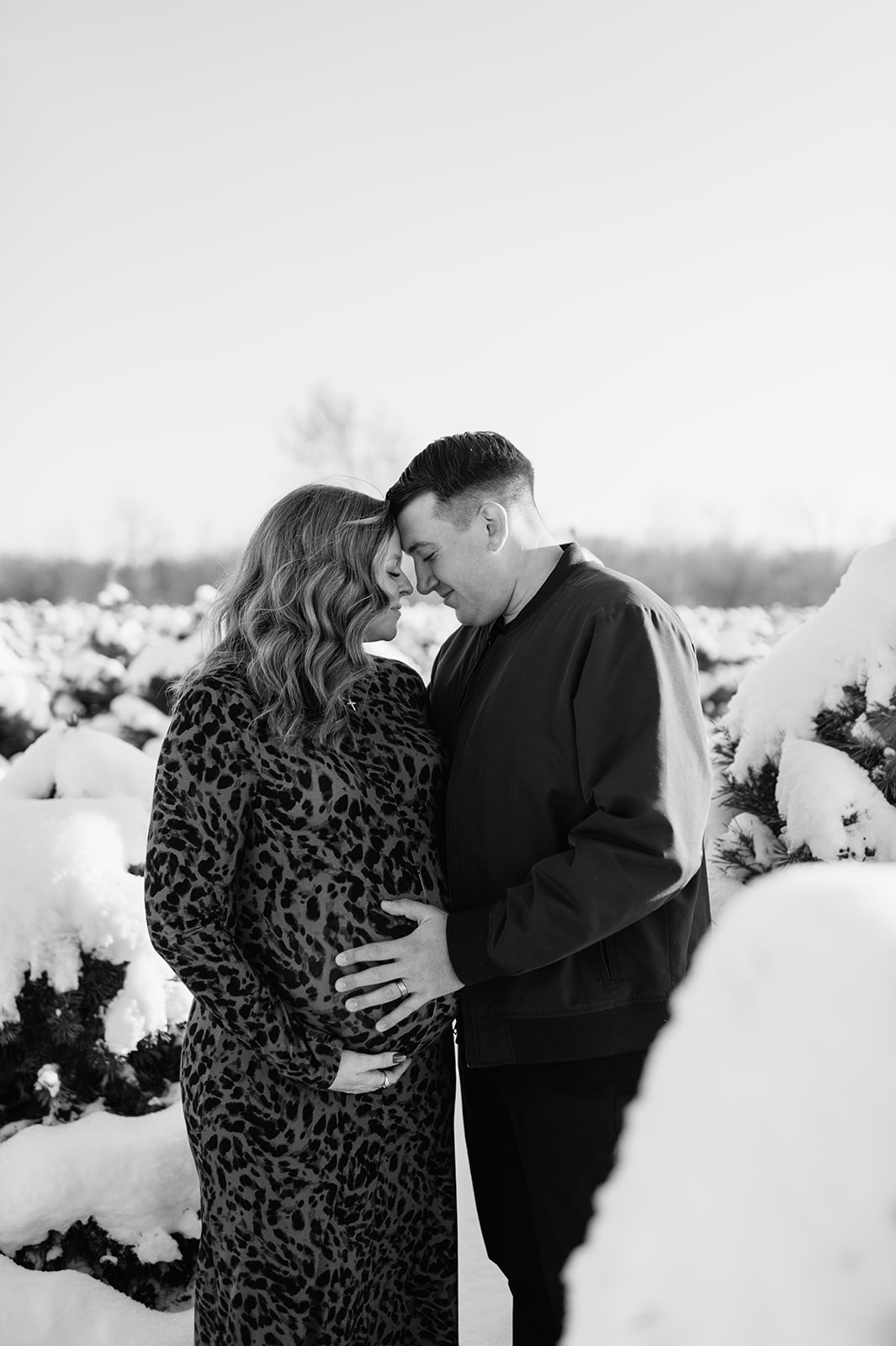 Black and white photo of a couple touching foreheads during their outdoor winter maternity photos in Northern Indiana at a tree farm.