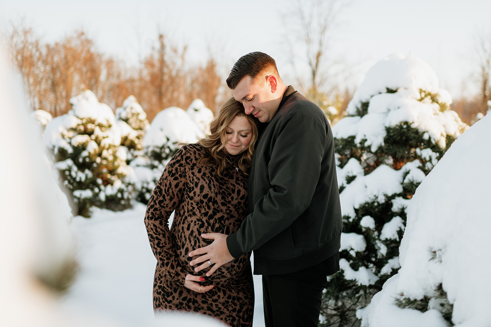 Expectant mother leaning on partner among snow-covered pine trees during outdoor winter maternity photos.