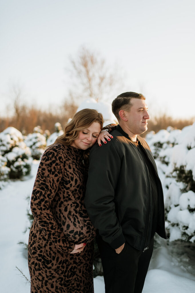 Expectant mother leaning on partner among snow-covered pine trees during outdoor winter maternity photos.