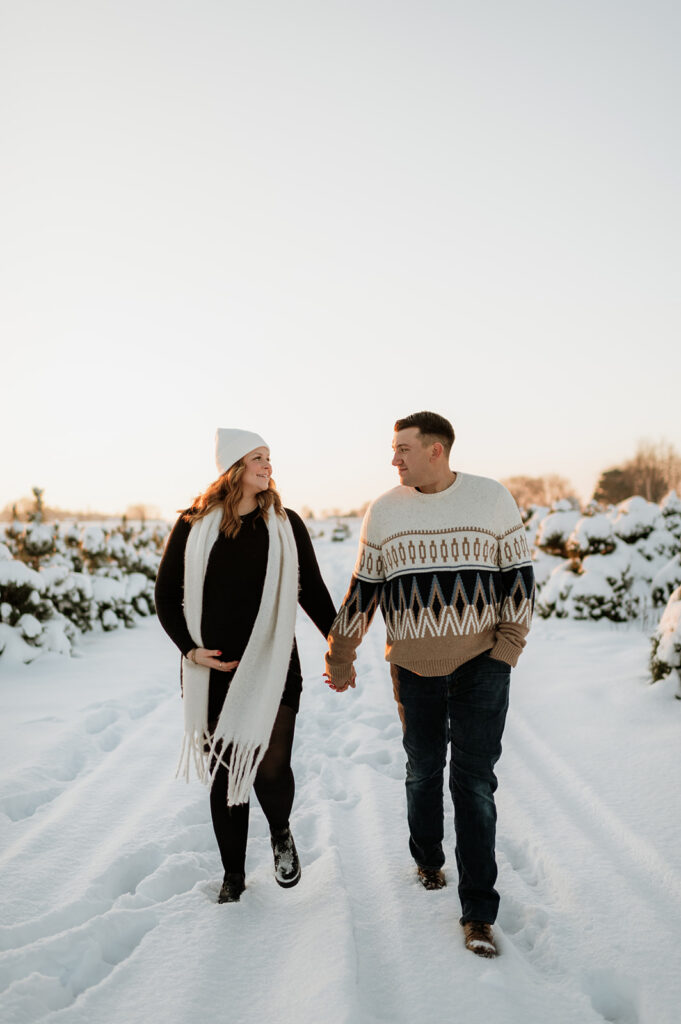 Couple walking through fresh snow between rows of pine trees during outdoor winter maternity photos at Flickinger Farms.