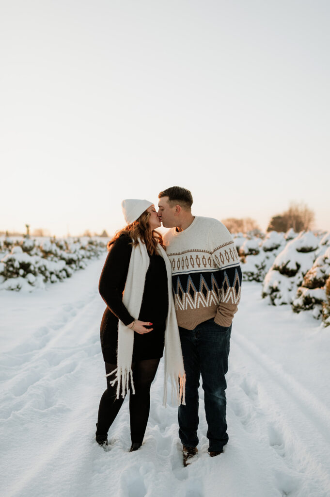 Couple sharing a kiss during golden hour winter maternity photos at a snowy tree farm in Elkhart, Indiana.
