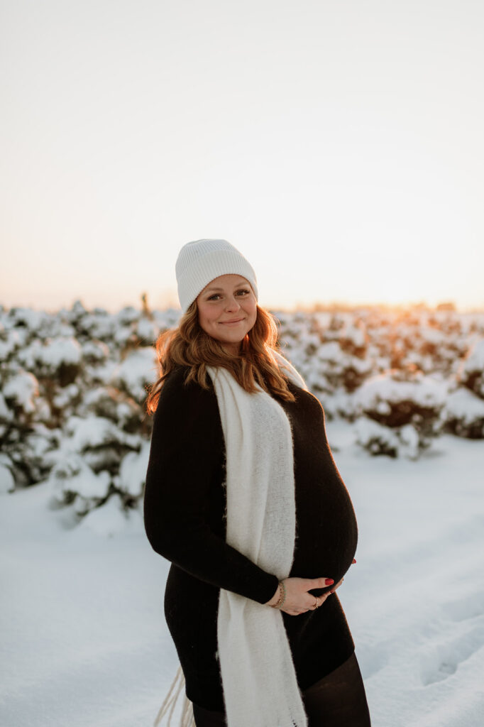 Pregnant woman standing in the snow surrounded by evergreen trees during outdoor winter maternity photos.