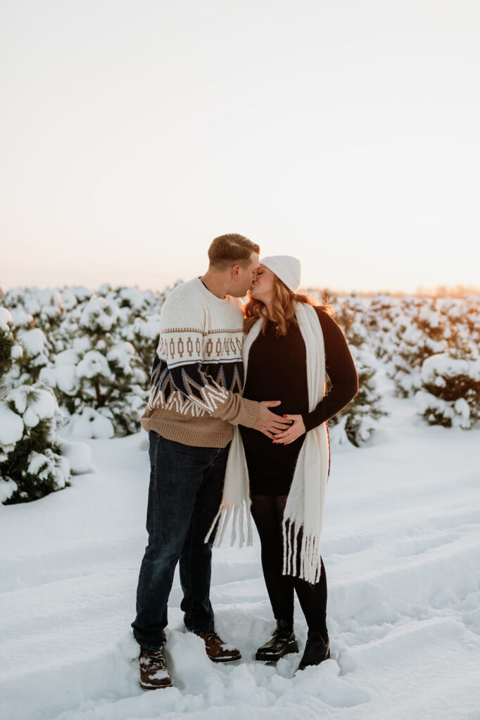 Expectant couple kissing while holding baby bump during sunset tree farm maternity photos at Flickinger Farms in Elkhart, Indiana.
