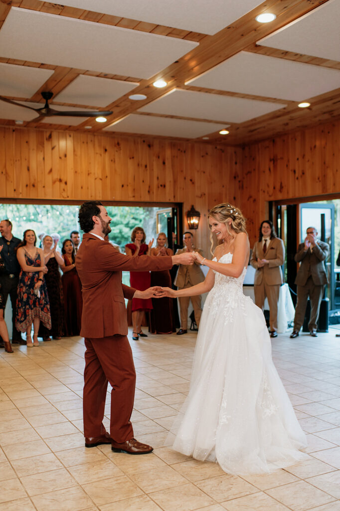 Bride and groom sharing their first dance surrounded by guests inside a wood-paneled reception pavilion.
