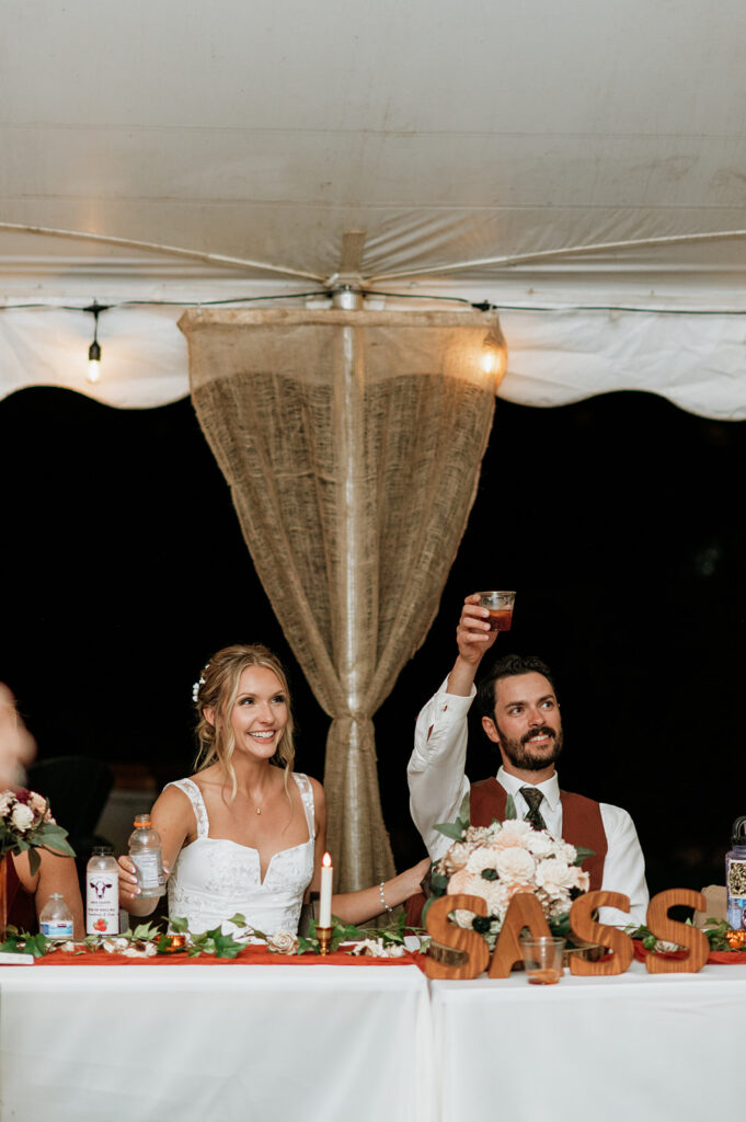 Bride and groom raising drinks during wedding reception toasts at their sweetheart table.