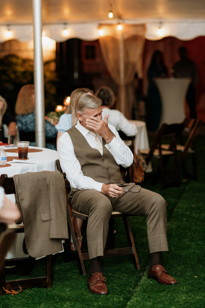 Father of the bride getting emotional and wiping away tears while listening to emotional reception speeches under a tent.