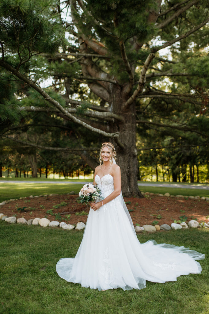 Bride standing with bouquet under a large pine tree at The Lodge at Rush Lakes wedding venue in La Porte Indiana.