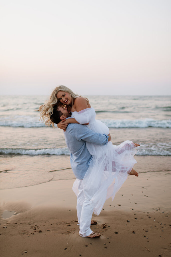 Man picking up his fiancé for their beach engagement photos at New Buffalo Beach during the summer.