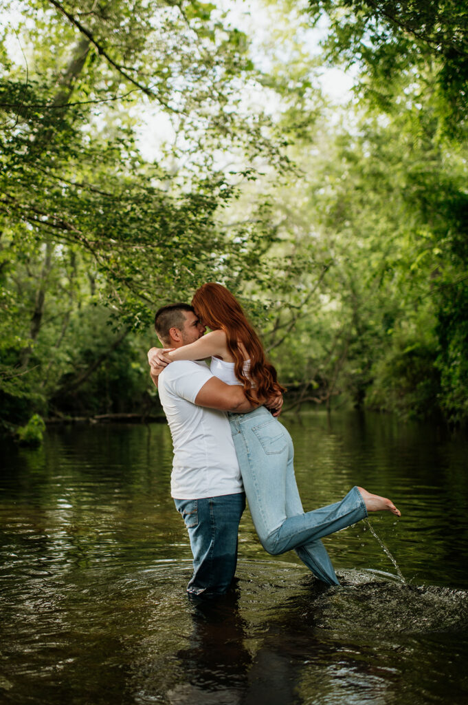 Man lifting up his fiancé during their casual summer engagement photos in a creek.