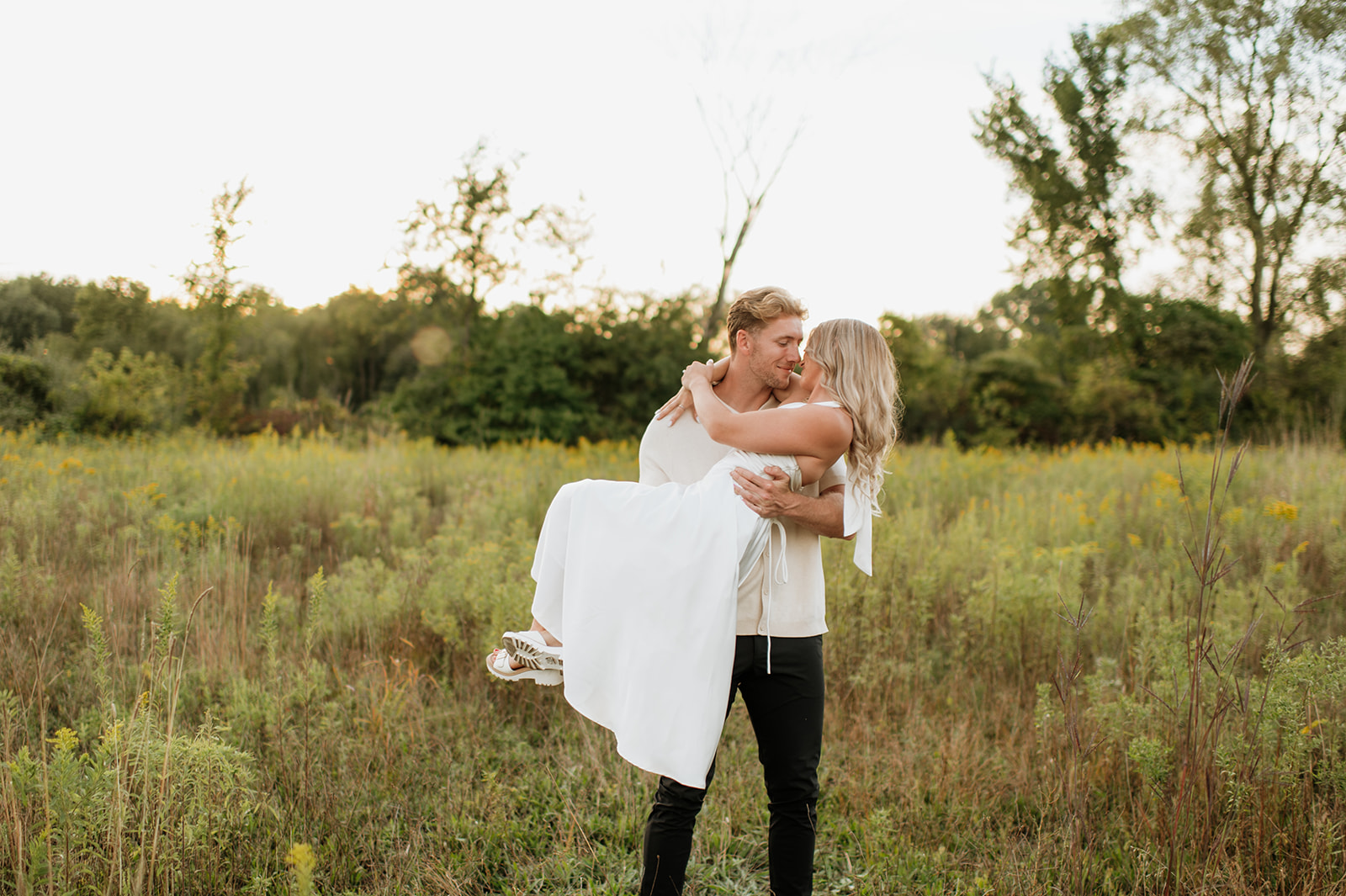 Man lifting his fiancé in his arms during their fall engagement photos in an open field in Goshen, Indiana.