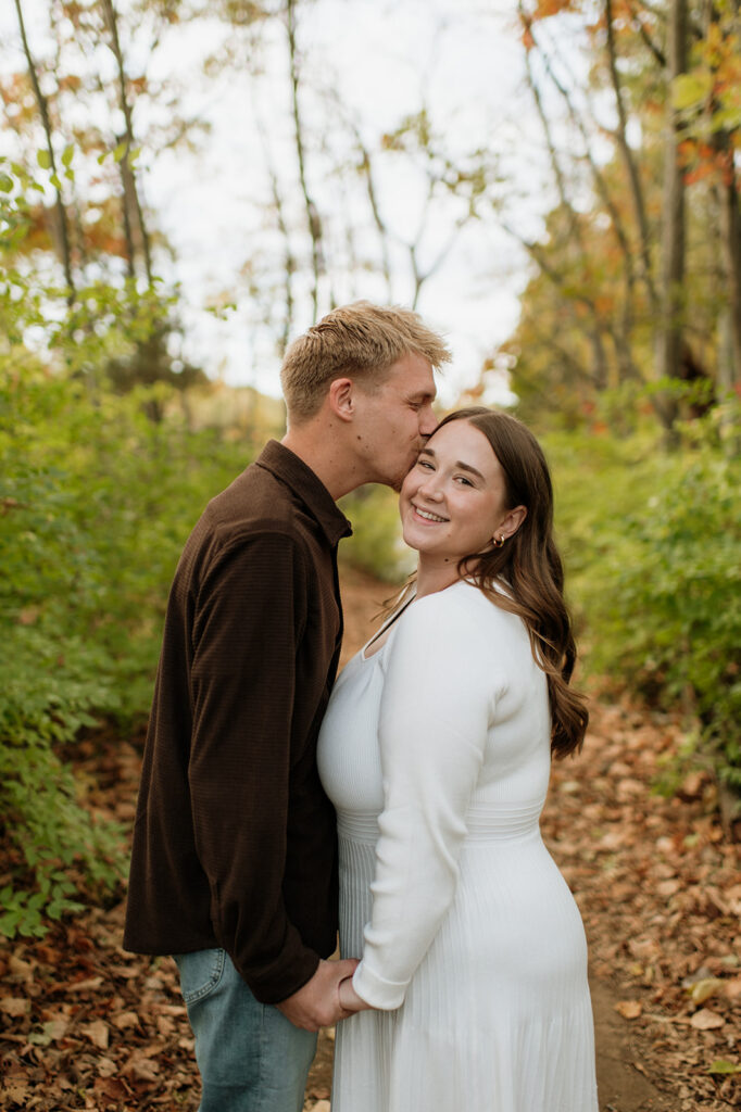 Man kissing his fiancé on the cheek during their outdoor fall engagement session.