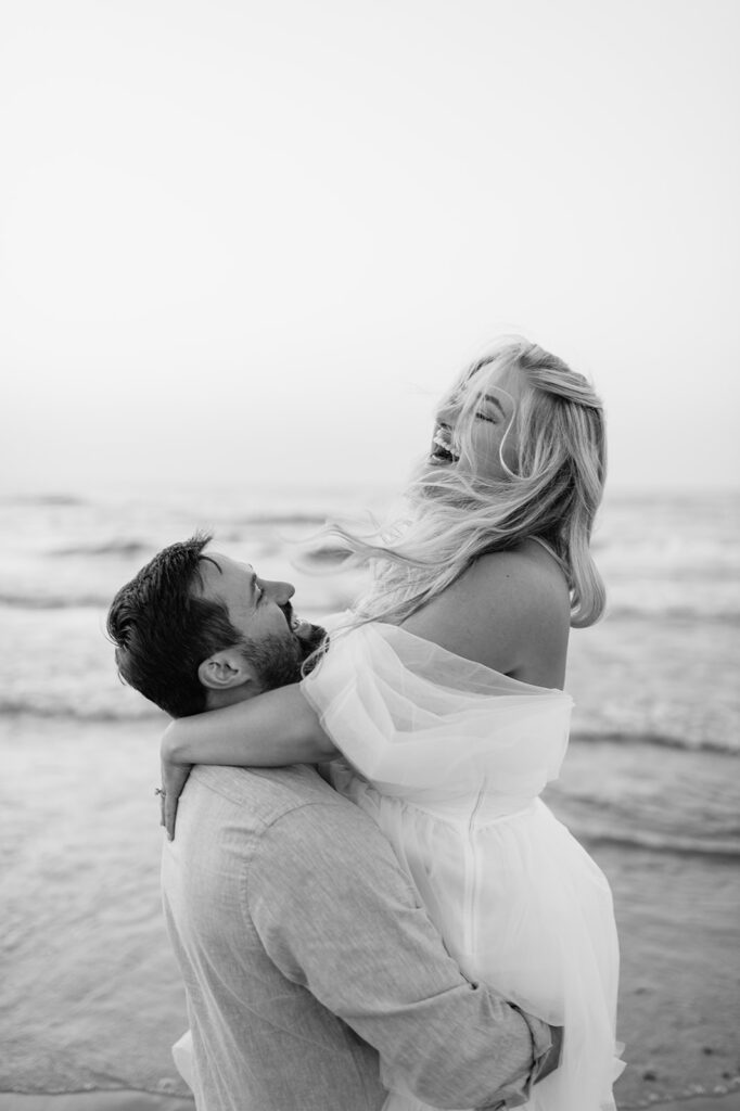 Candid black and white photo of a man lifting up his fiancé during their New Buffalo Beach engagement photos.