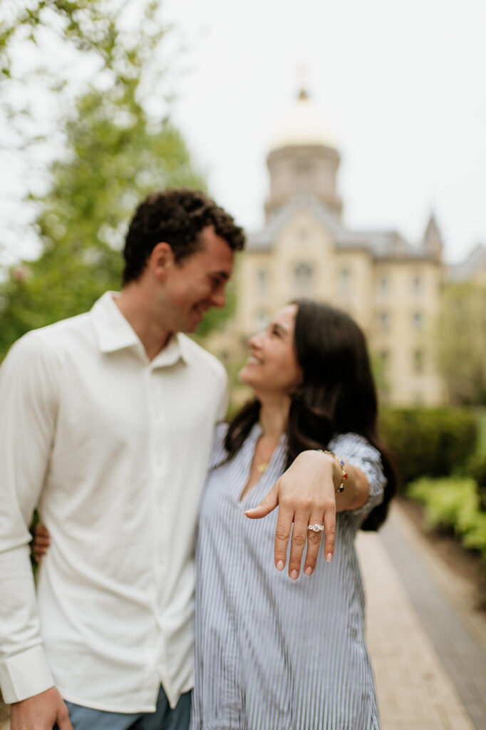 Couple standing together in front of the iconic Golden Dome while the woman shows off her engagement ring after a surprise Notre Dame proposal.