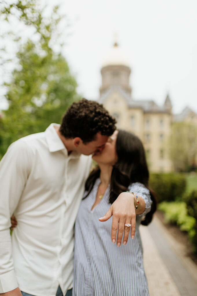 Man and woman kissing while the woman shows off her engagement ring after their Notre Dame proposal in front of the Golden Dome.
