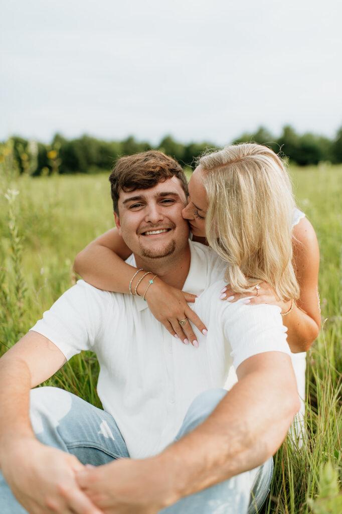 Woman kissing her fiancé on the cheek as they sit in a grassy field for their Cobus Creek County Park Engagement Photos in Elkhart, Indiana.