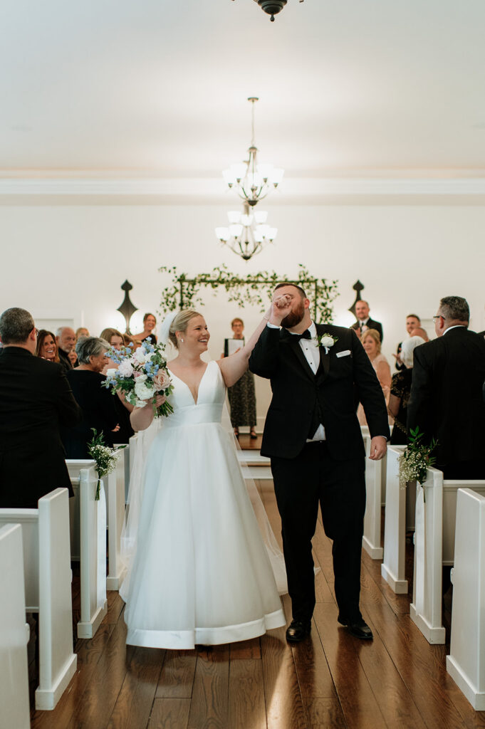 Groom kissing his brides hand as they walk back down the aisle as husband and wife after their Morris Estate Chapel wedding ceremony.